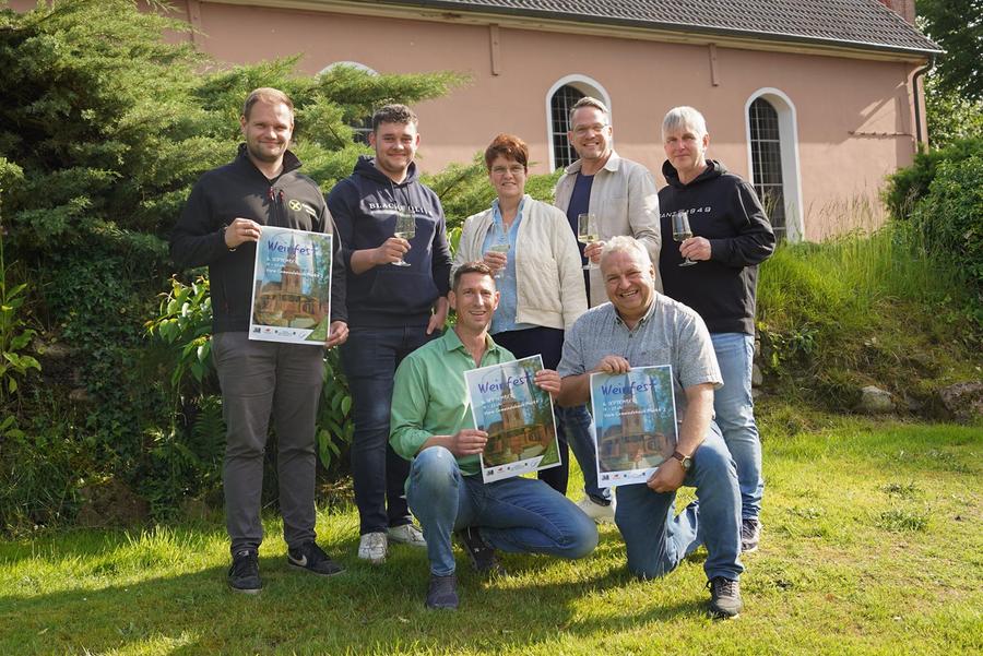 Sieben Personen stehen und knien auf einer sonnigen Wiese vor der Kirche in Großenkneten. Alle halten Weingläser in der Hand, drei von ihnen zudem Plakate mit Werbung für das Weinfest Großenkneten am 6. September 2025. Im Hintergrund sind grüne Büsche und die rosa Fassade der Kirche zu sehen. Die Gruppe wirkt fröhlich und einladend.